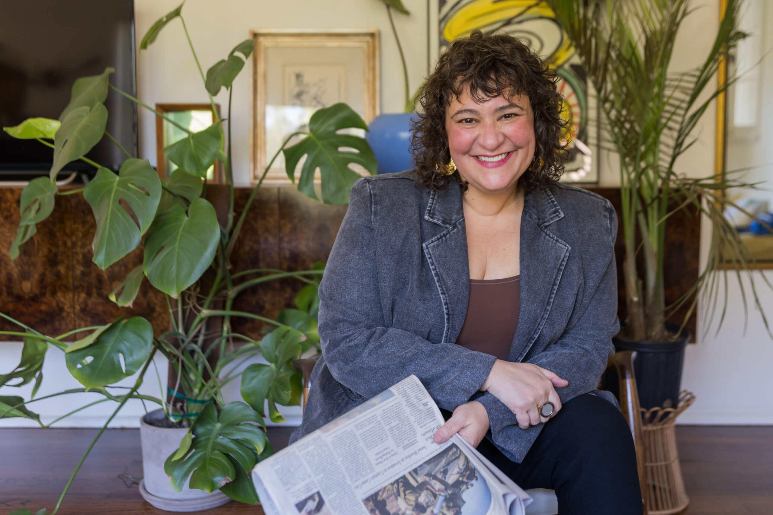 Person smiling while seated indoors holding a newspaper, in a cozy, plant-filled space, during a casual coffee chat conversation.